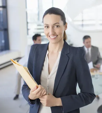 Healthcare administrator smiling in a professional office setting while holding a folder, with a team discussing in the background.